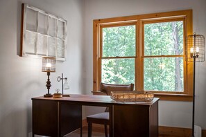 A room with a wooden desk, a lamp, a chair, a window with a view of trees, and a wall-mounted shelf with a glass container. - Maison Bleue - Close to Downtown | Game Room | Firepit (Blue Ridge)