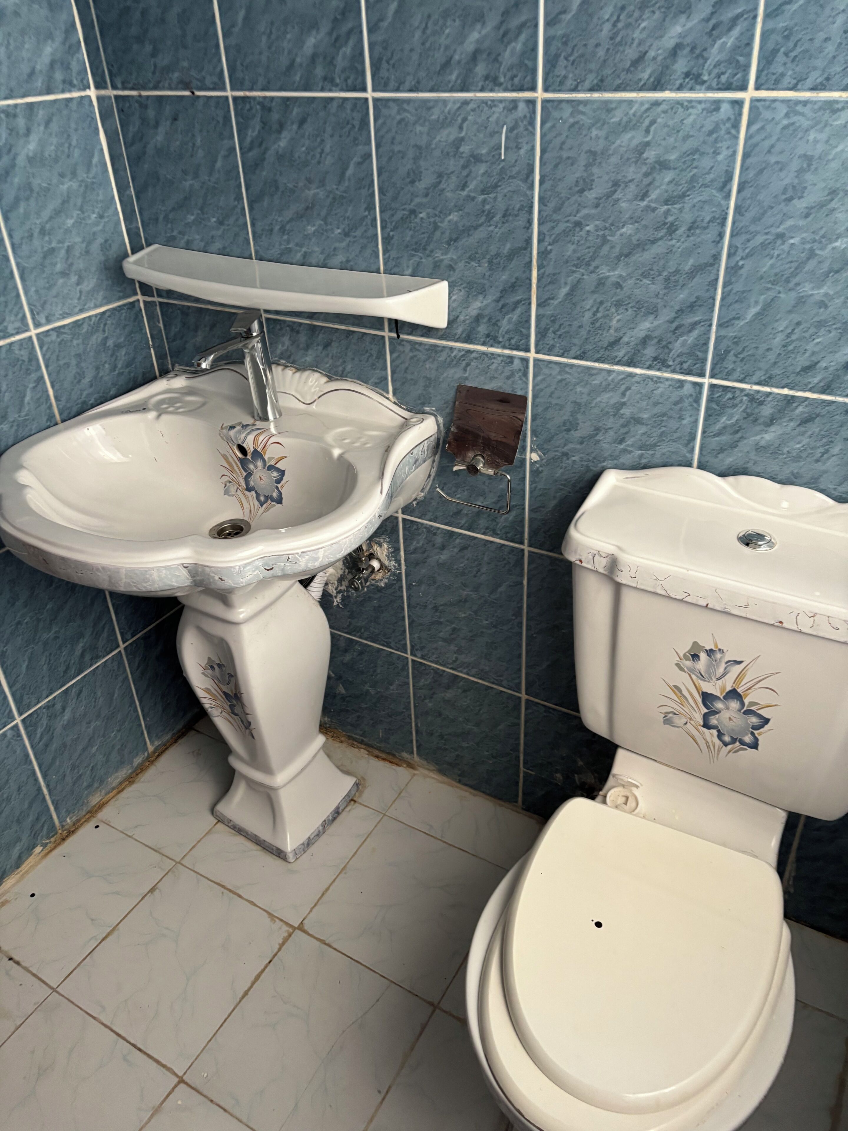 Un baño con lavamanos e inodoro, paredes con azulejos azules y suelo de baldosas.