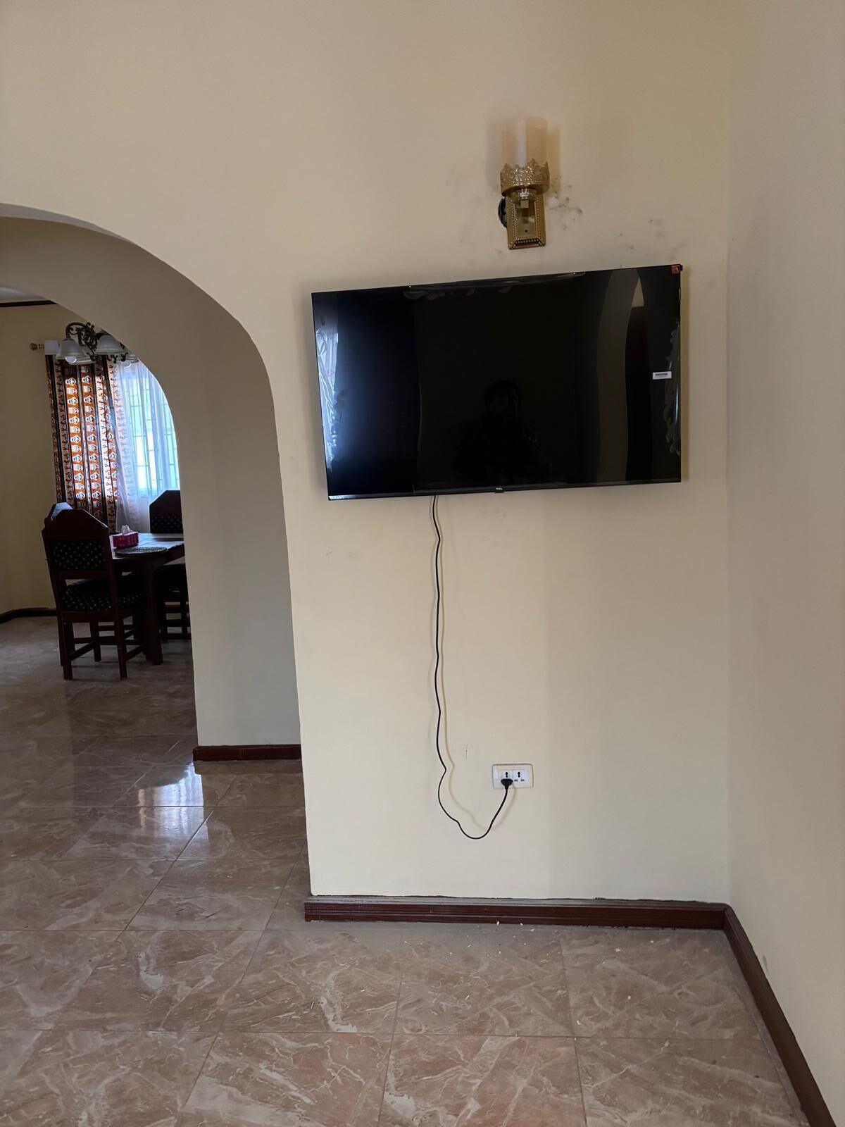 Una sala de estar con televisor de pantalla plana montado en la pared, un comedor con sillas y una mesa, y piso de baldosas.