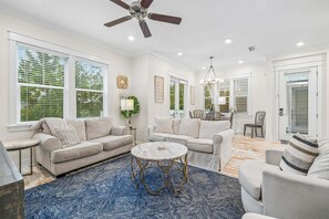 A spacious living room with a ceiling fan, large windows with blinds, a round coffee table, and a blue patterned rug. - Prominence, LSV/Golf Cart & Bikes! Easy Walk to Pool/The Big Chill! (Panama City)