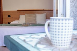 Ein ordentlich bezogenes Bett mit Kissen und einer grünen Decke, ein Nachttisch mit einer weißen Tasse und ein Fenster mit Blick ins Freie.