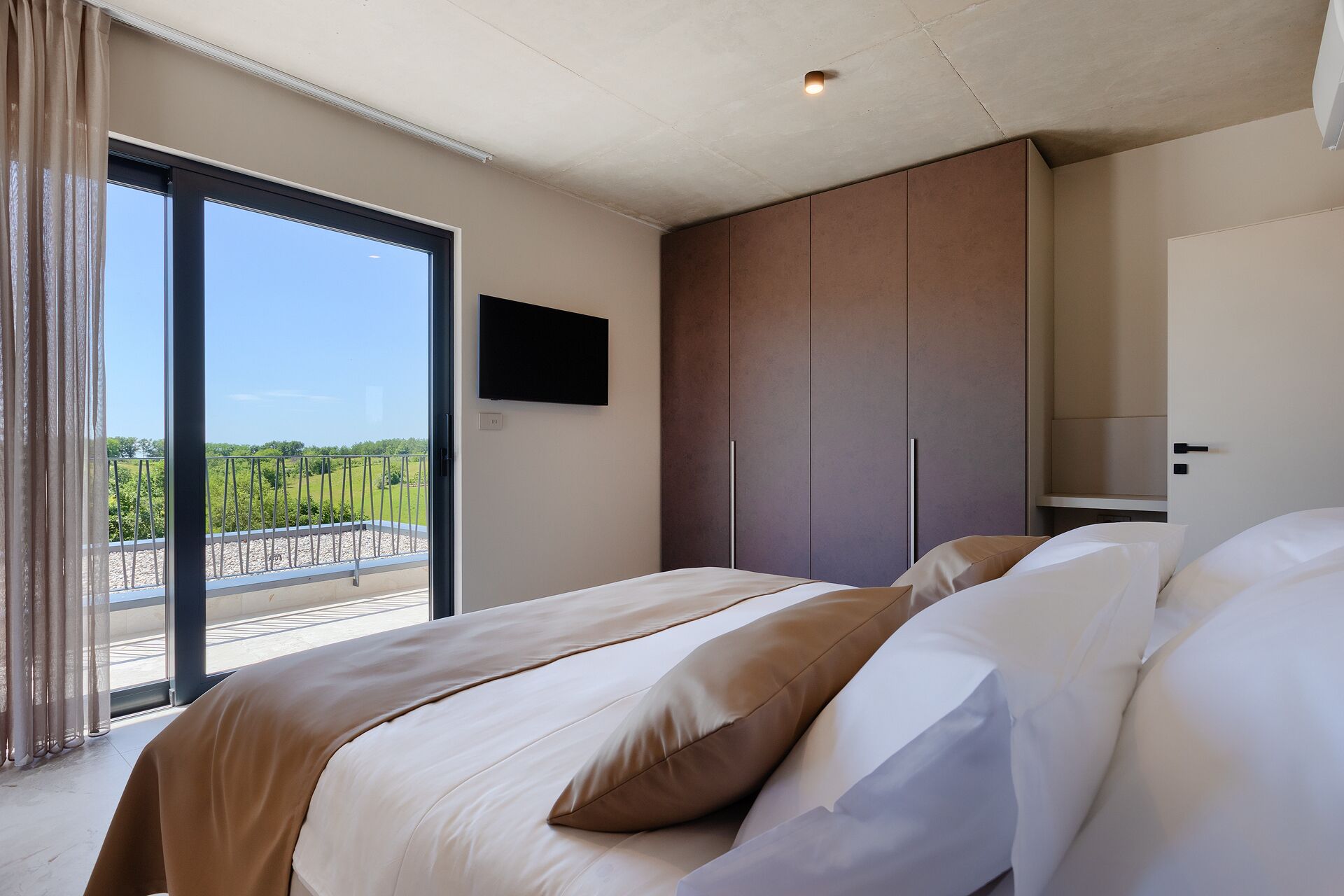 Un dormitorio con cama, televisor montado en la pared y vistas a zonas verdes por un amplio ventanal.
