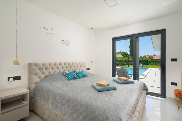 Un dormitorio con una cama grande, una mesita de noche, una lámpara de pared y un ventanal corredero a un espacio exterior.