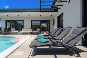 Un área de piscina moderna al aire libre con sillones de descanso, un porche con paredes de vidrio y un cielo azul despejado.