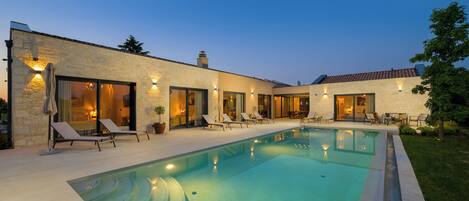 Una casa moderna con piscina, sillas de descanso y un patio bien iluminado.