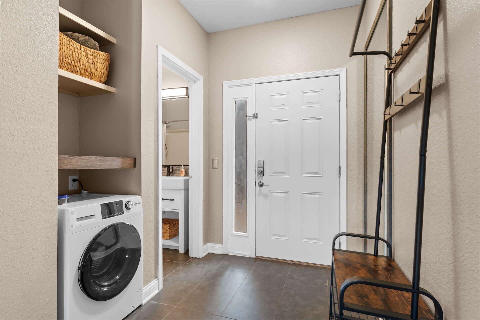 A laundry room with a washing machine, shelves with baskets, a door, and a chair.
