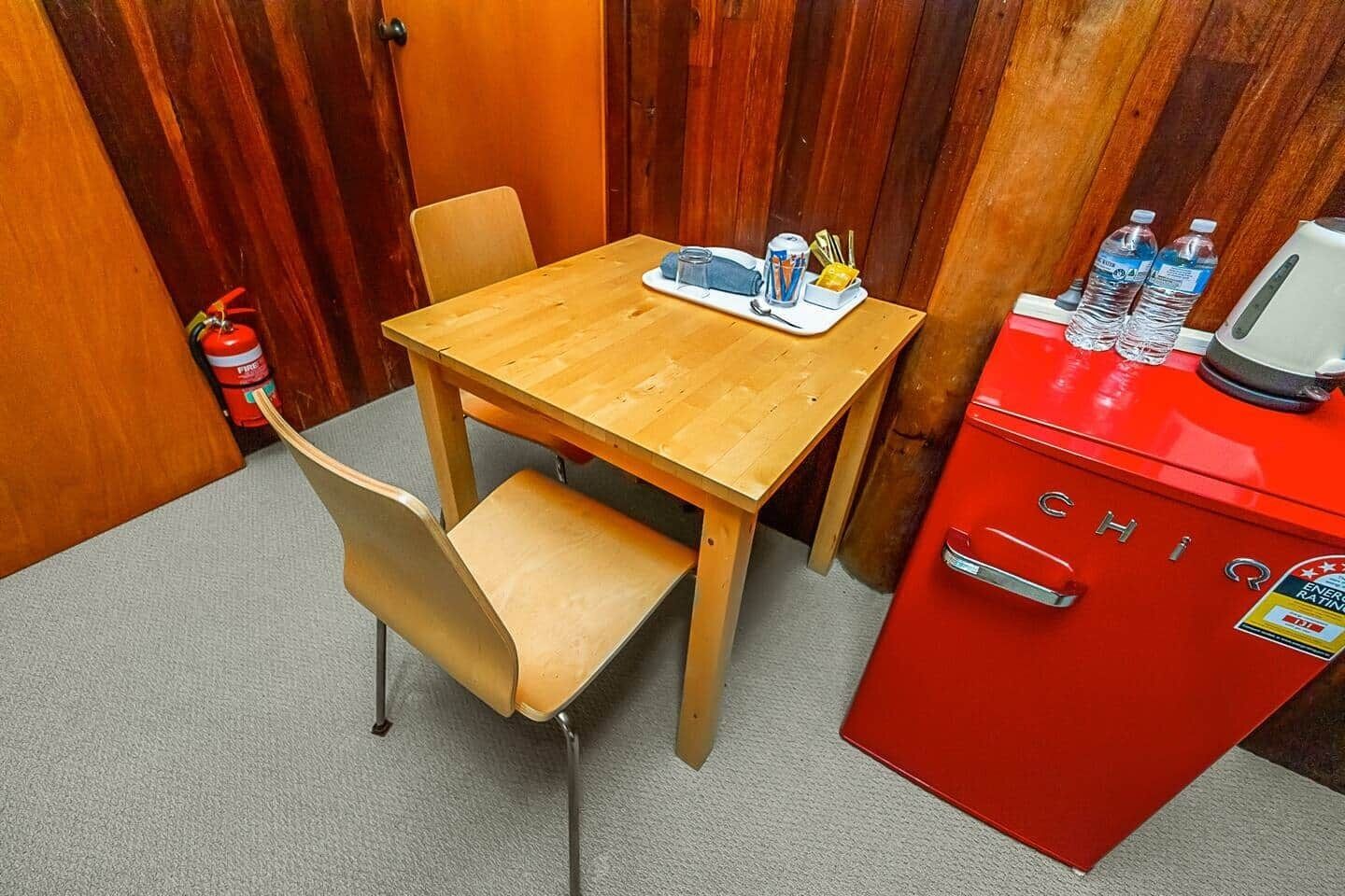 Una habitación pequeña con una mesa de madera, dos sillas, un armario rojo que contiene una botella de agua y una lata, y un extintor.