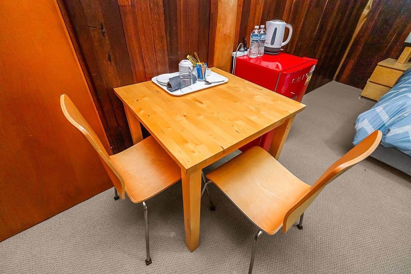 Una pequeña mesa de madera con dos sillas, un armario rojo y una cama con una colcha azul a cuadros.
