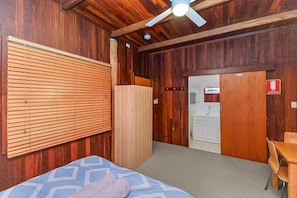 Una habitación con una cama, un ventilador de techo, un armario de madera y una kitchenette al fondo.