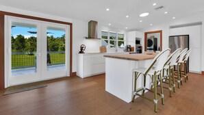 A modern kitchen with white cabinetry, a central island, and a view of a green landscape through large sliding doors. - Bleu Hermitage | Private 25-Acre Luxury Sanctuary (Naples)