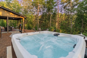 Ein Whirlpool umgeben von einer Holzterrasse und Stühlen, mit einer überdachten Sitzecke und Blick auf einen Wald.