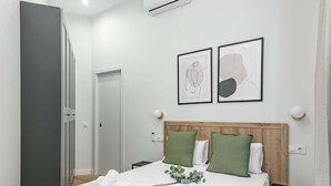 Una camera d'albergo con un letto, un comodino, un tavolo da notte, un condizionatore d'aria a muro e due opere d'arte astratte incorniciate.