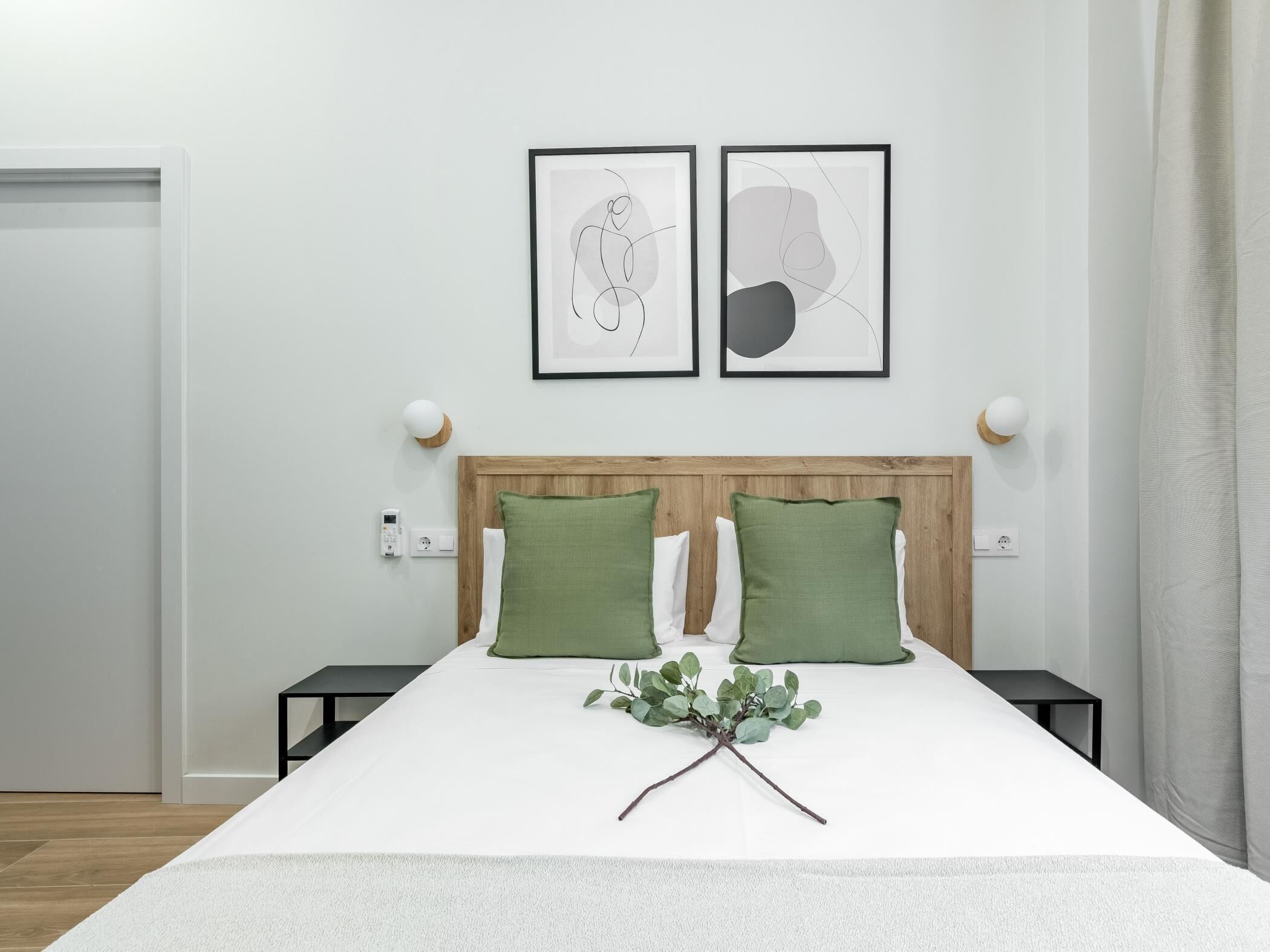 Un dormitorio ordenado con cabecera de madera, dos almohadas verdes, una mesita de noche con lámpara y dos cuadros abstractos enmarcados en la pared.