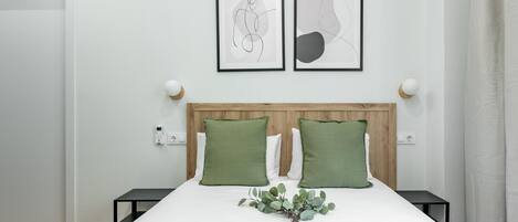 A neatly arranged bedroom with a wooden headboard, two green pillows, a bedside table with a lamp, and two framed abstract artworks on the wall.