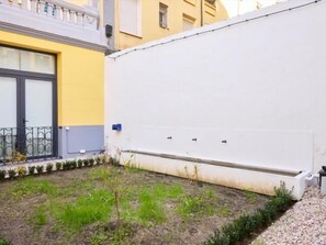 Un patio con un muro blanco, un pequeño jardín y un edificio de fachada amarilla.