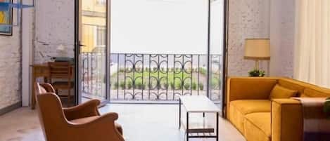 A room with a sofa, a chair, a small table, and a balcony with a view.