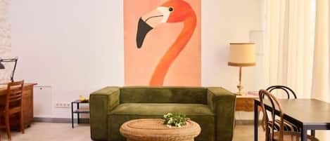 A living room with a green sofa, a wicker coffee table, a dining table with chairs, and a large painting of a flamingo on the wall.