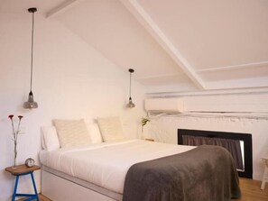 Ein Schlafzimmer mit einem Bett, zwei Pendelleuchten, einem Nachttisch mit einer Uhr und einem Fenster mit Vorhängen.