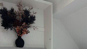 Ein ordentlich bezogenes Bett mit einem flauschigen Kopfkissen und einem Stapel weißer Handtücher, eine Vase mit getrockneten Blumen und ein Laptop auf einem Schreibtisch in einem Raum mit geneigter Decke.
