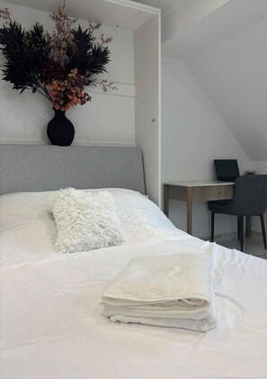 A neatly made bed with a fluffy pillow and a stack of white towels, a vase with dried flowers, and a laptop on a desk in a room with a sloped ceiling. - Lavish, The White Apartment (Bury St Edmunds)