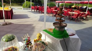 A dining area with outdoor tables and chairs, a fountain centerpiece, and a variety of food items on display. - Park (Bihac)