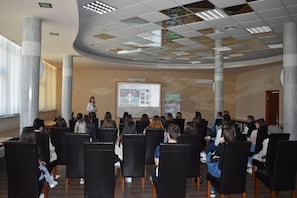 A conference room with rows of chairs facing a presentation screen, large columns, and a high ceiling with recessed lighting.