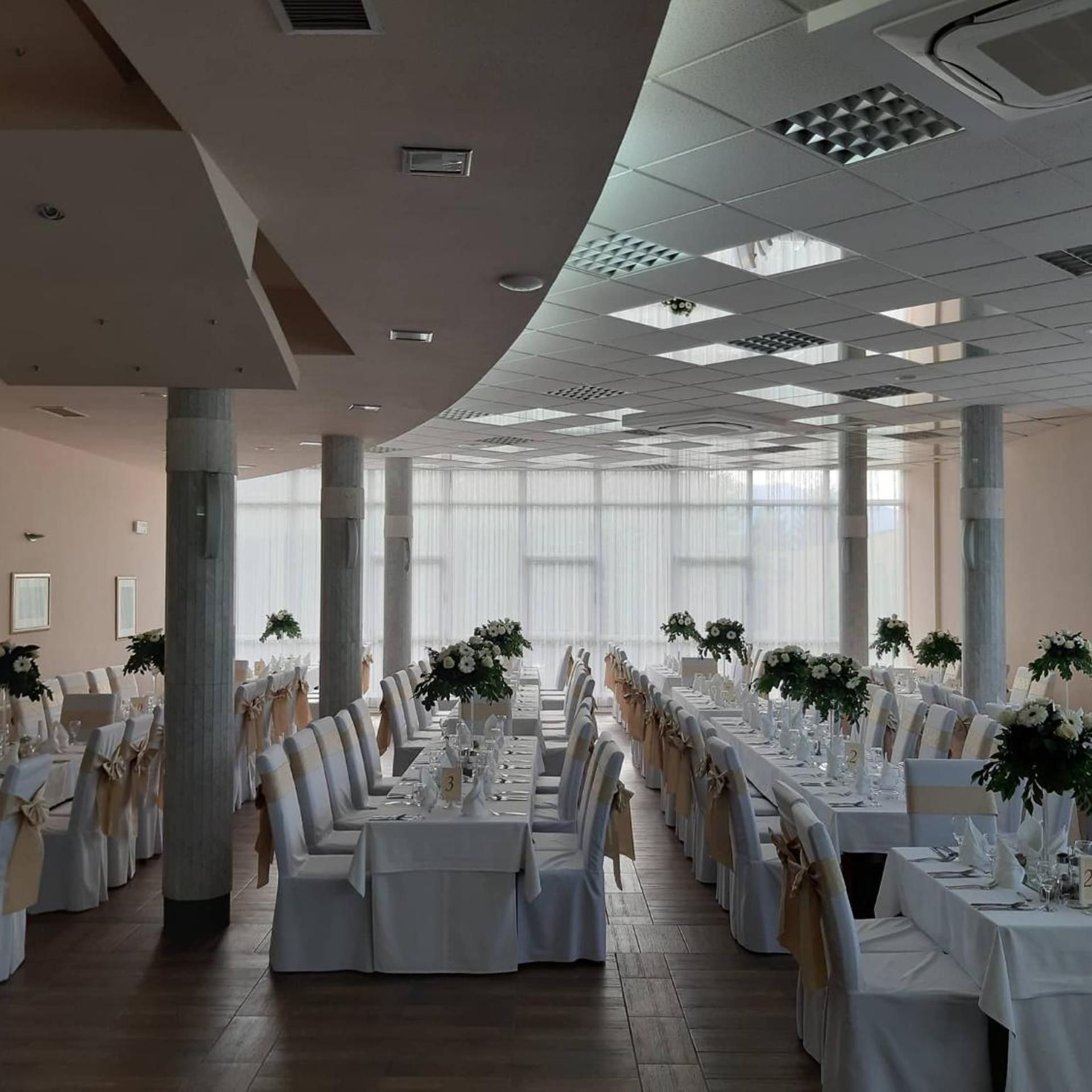 A spacious banquet hall with high ceilings, large windows, and rows of tables set for a formal event.