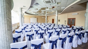 Una sala de conferències amb files de cadires blanques amb llaços blaus, un sostre corbat amb il·luminació empotrada i una pissarra gran.