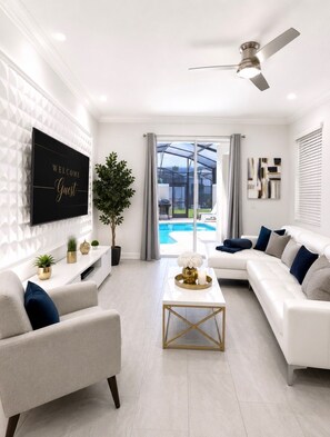 A modern living room with a white sofa, a coffee table, a TV, and a view of a swimming pool through glass doors. - Spacious 4-bedroom house with AC in pleasant Kissimmee (Kissimmee)