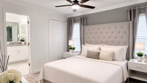 Een slaapkamer met een groot bed, een plafondventilator, een nachtkastje met een lamp en een dressoir met een schaal decoratieve objecten.