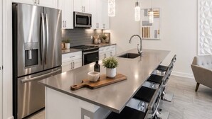Een moderne keuken met roestvrijstalen apparatuur, een centraal eiland met barkrukken, hanglampen en een wandkunstwerk met geometrische vormen.