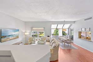 A modern living room with a flat-screen TV, a sofa, a dining table, and a large window with curtains. - Shelter Cove Villa, Lagoon Views, Community Pool, Cozy Furnishings! (Hilton Head Island)