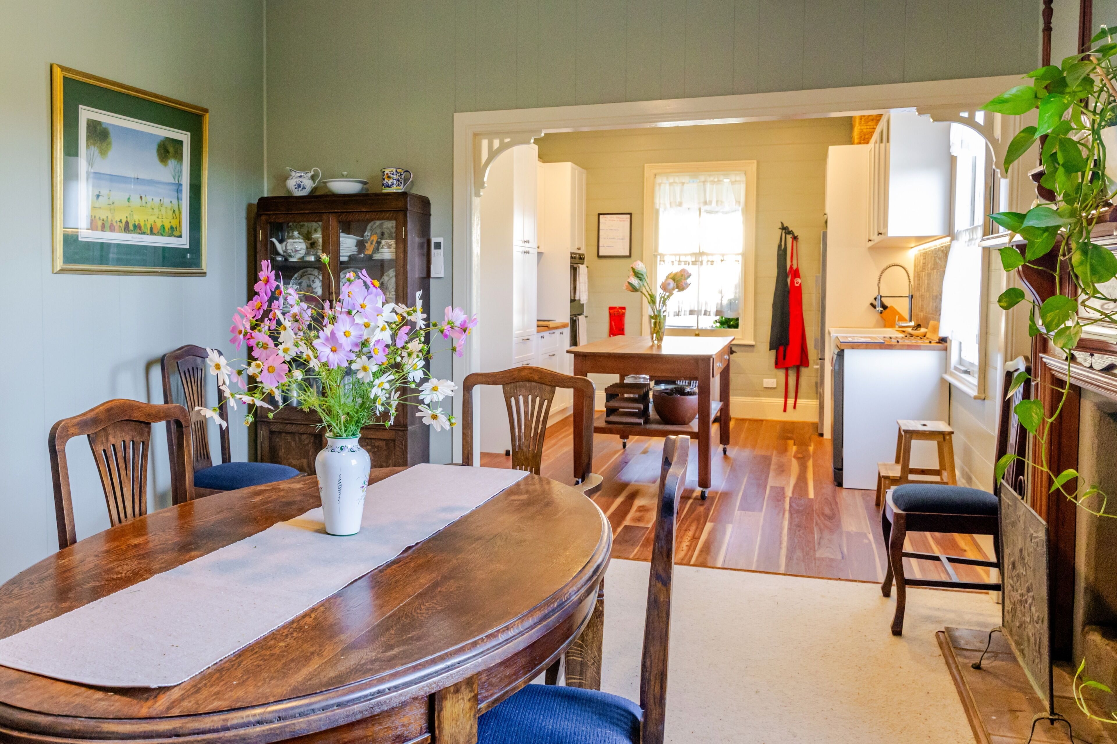 A dining area with a wooden table, chairs, a vase of flowers, and a kitchen in the background.