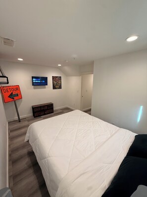 A bedroom with a bed, a television mounted on the wall, a small cabinet, and a door leading to another room.
