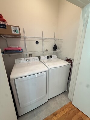 A laundry room with two white front-loading washers, a metal shelf with folded towels, and a cardboard box on the left.
