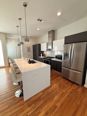 A modern kitchen with a central island, stainless steel appliances, pendant lighting, and a wooden floor.