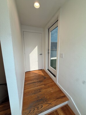 Un couloir avec un plancher en bois, une porte blanche et une fenêtre donnant sur l’extérieur.