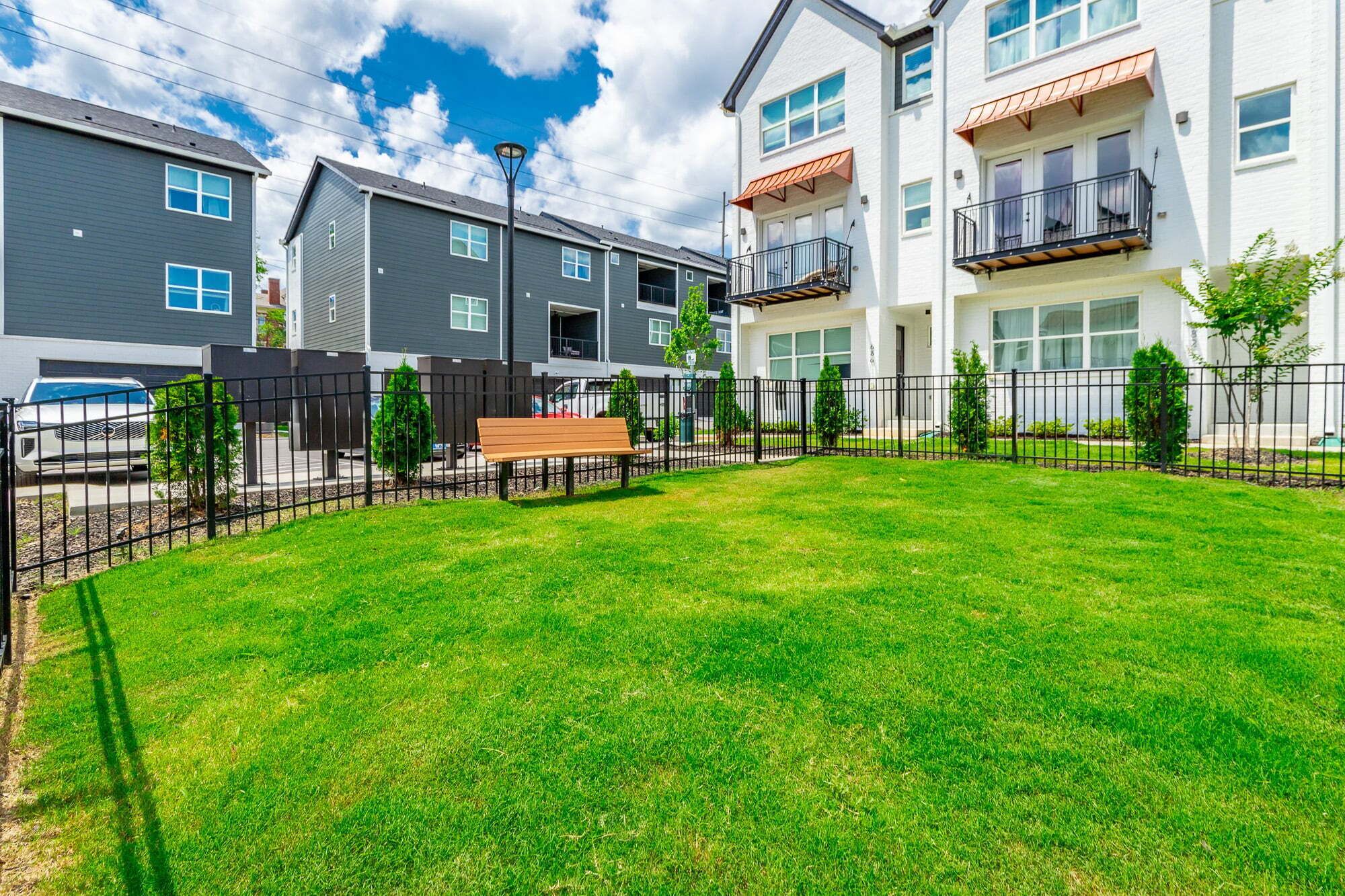 A residential complex with multiple apartment buildings, a fenced yard with a bench, and a street with parked cars.
