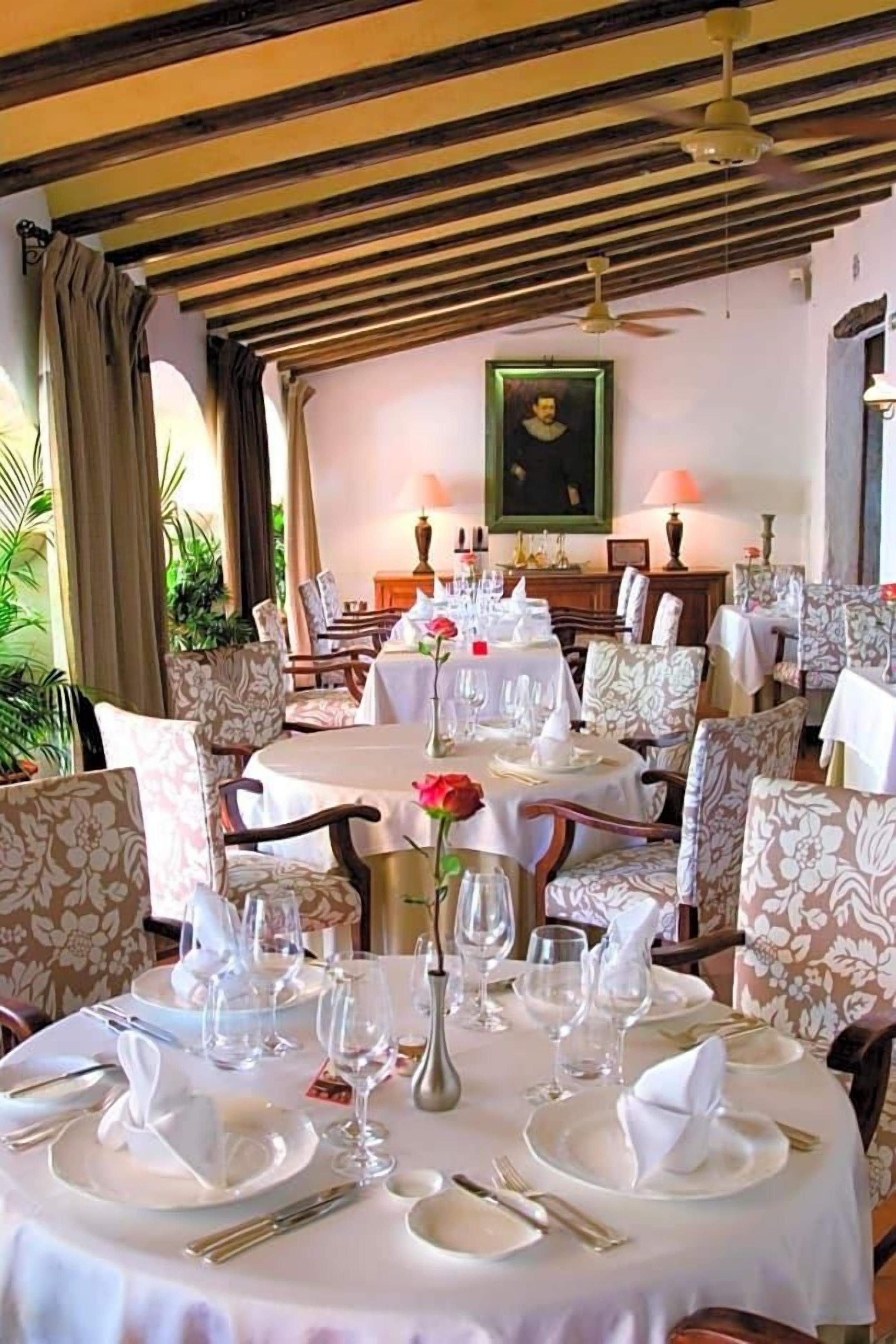 A dining area with round tables set for a meal, chairs with floral upholstery, and a ceiling fan.