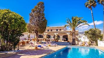 Un área junto a la piscina con sillones de descanso, piscina y un edificio con ventanas de arco y techo de tejas.
