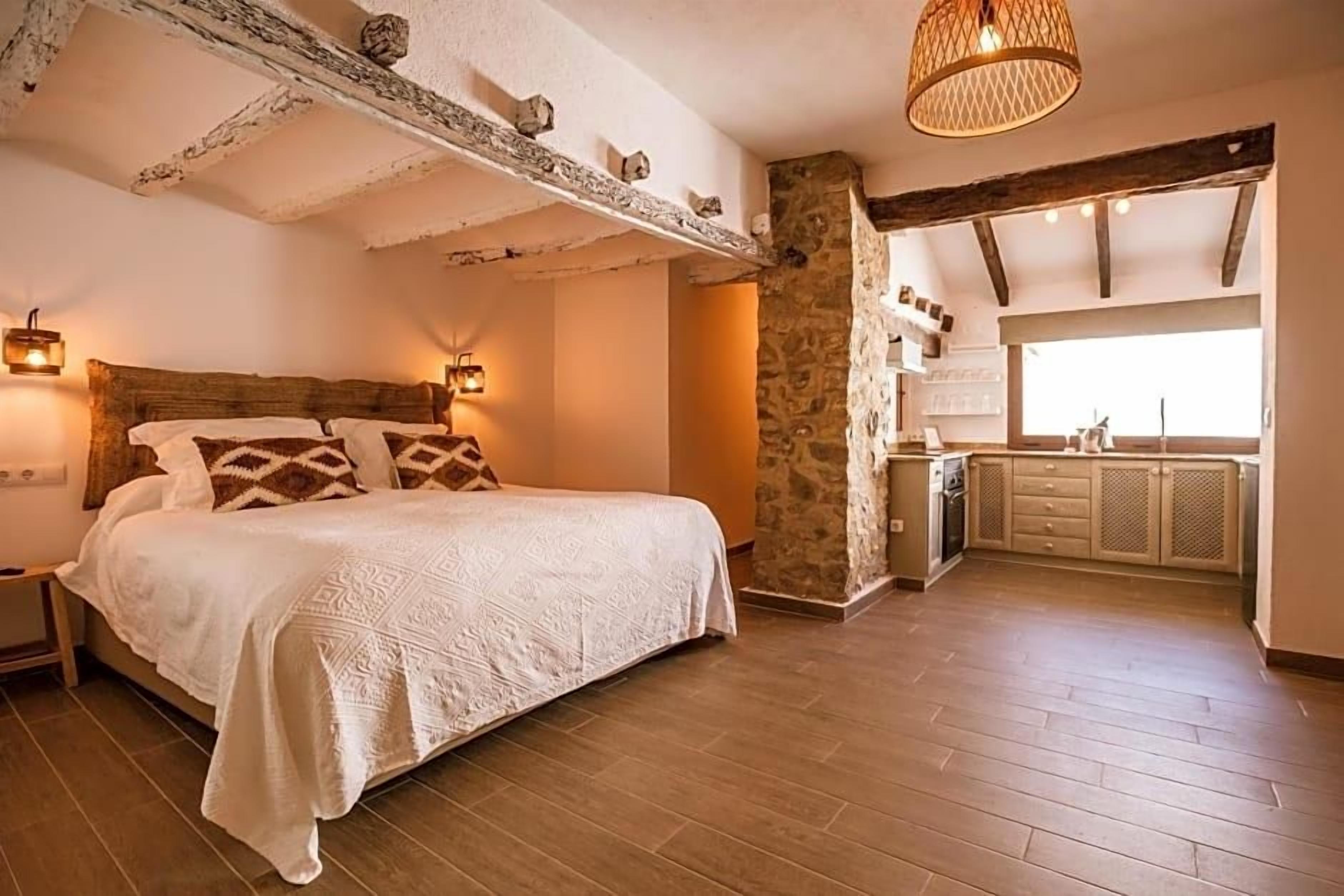 A bedroom with a large bed, a stone wall, and a kitchen area visible through an open doorway.