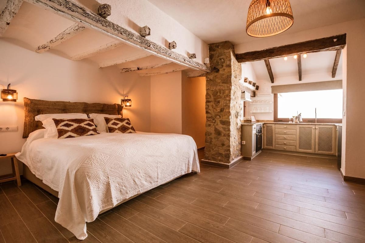 Ein Schlafzimmer mit einem großen Bett, einer Steinwand, einer Holzbalkendecke und einer Küchenzeile mit Schränken und einem Fenster.
