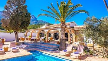 Un área junto a la piscina con sillones de descanso, una palmera y un edificio con arcos.