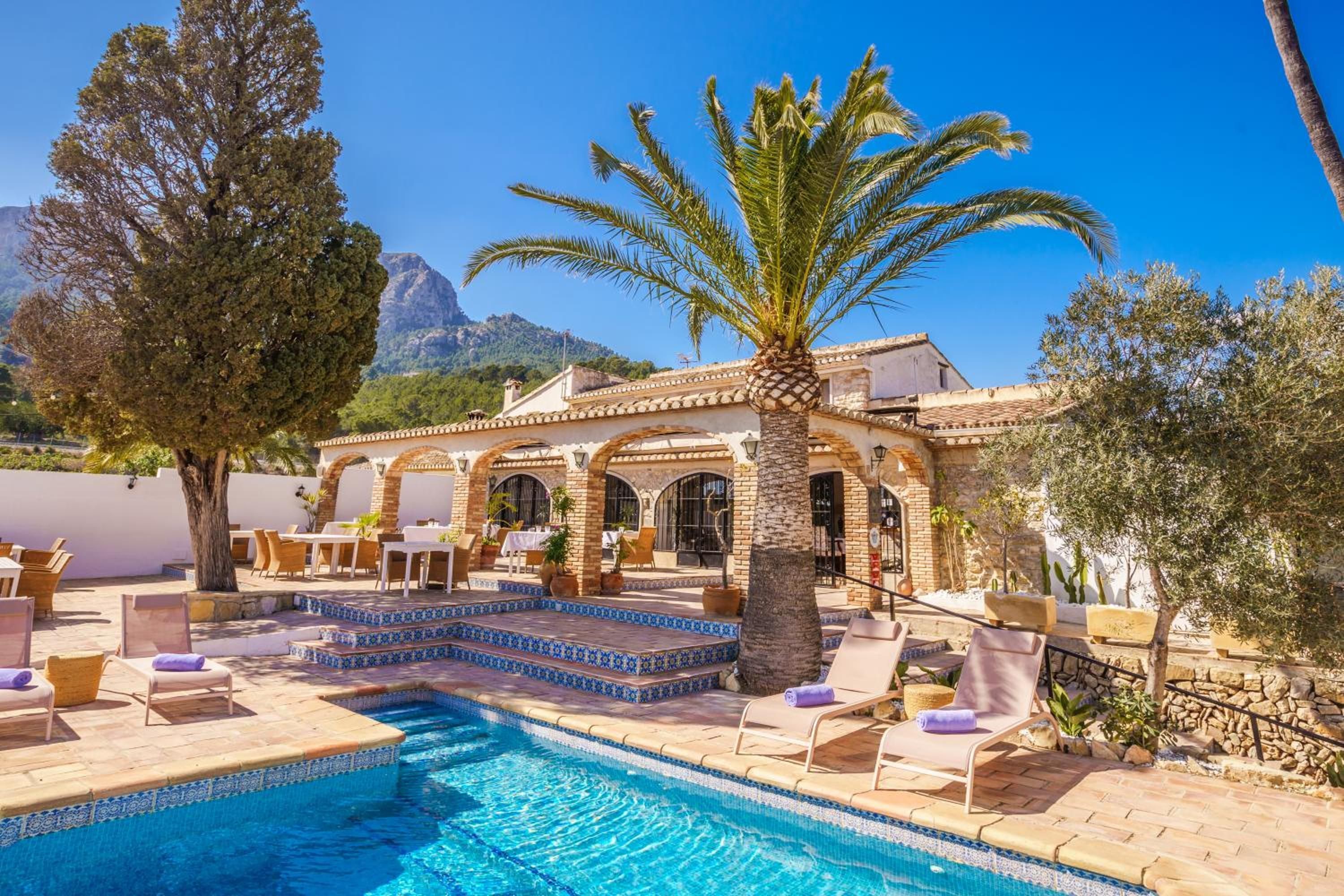 Zona de piscina con sillones, una palmera y un edificio con arcos.