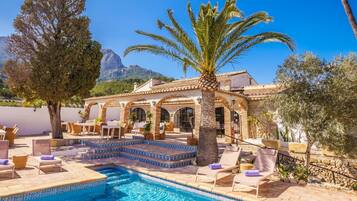 Zona de piscina con sillones, una palmera y un edificio con arcos.