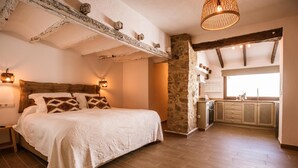 A bedroom with a large bed, a stone wall, a wooden ceiling, and a kitchen area with cabinets and a window. - Hotel Restaurante Casa Del Maco (Benissa)