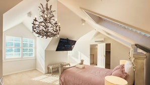 A bedroom with a sloped ceiling, a chandelier, a bed with a pink bedspread, a bedside table, and a television mounted on the wall.