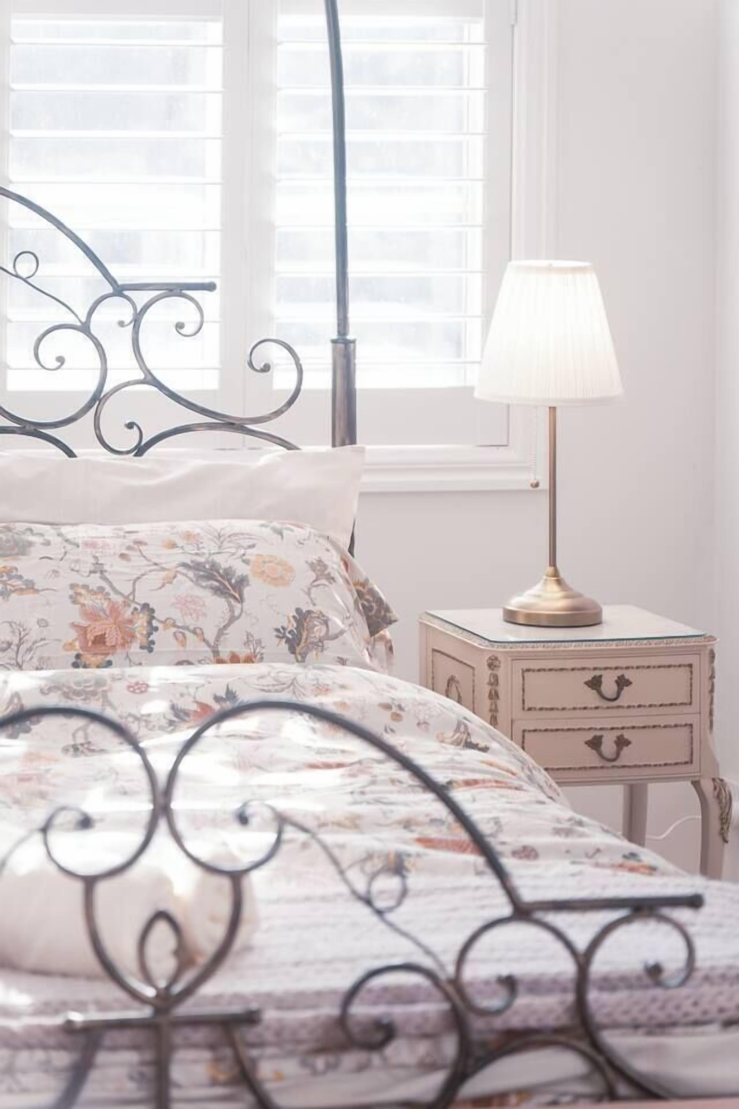Un lit avec une couvre-lit à motifs floraux, une tête de lit en métal, une table de chevet avec une lampe et une fenêtre avec des stores.