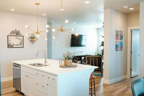 Una cocina moderna con una isla central, iluminación colgante y un área de comedor con una silla.