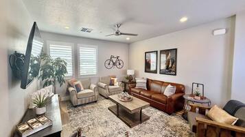 A living room with a sofa, armchairs, a coffee table, and a television. There are two windows with blinds, a ceiling fan, and wall art.